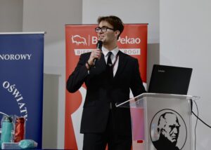 Na zdjęciu stoi młody mężczyzna w eleganckim czarnym garniturze, białej koszuli i czarnym krawacie. Ma krótkie brązowe włosy, okulary i trzyma mikrofon w prawej dłoni, mówiąc do publiczności. Jego twarz jest uśmiechnięta i pełna pewności siebie.

Po lewej stronie znajduje się przezroczysta mównica z nadrukowanym czarnym wizerunkiem mężczyzny w profilu. Na mównicy stoi otwarty laptop marki Lenovo. W tle widać dwa roll-upy: niebieski oraz czerwony z charakterystycznym białym symbolem graficznym.

Scena wygląda jak prowadzenie konferencji lub moderowanie spotkania.