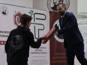 Nastolatek w czarnej bluzie stoi po lewej stronie kadru i odbiera nagrodę – długie prostokątne pudełko w czerwono-czarnych barwach – od dorosłego mężczyzny w granatowym garniturze. Mężczyzna ma krótkie, ciemne włosy i starannie przystrzyżoną brodę. Wręczając prezent, lekko się pochyla i uśmiecha.

W tle widać roll-upy z logotypami wydarzenia edukacyjnego, głównie w białej i zielonej kolorystyce. Zdjęcie dokumentuje oficjalny moment wręczenia nagrody uczestnikowi.