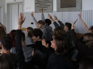 Zdjęcie przedstawia dużą grupę uczniów siedzących w sali. Widać głównie ich profile i tyły głów. Niemal wszyscy mają ręce uniesione wysoko do góry, zgłaszając się lub odpowiadając na pytanie prowadzącego. Uczniowie są w wieku nastoletnim, mają różne kolory włosów – blond, brązowe, ciemne – oraz ubrania w odcieniach szarości, czerni i niebieskiego.

Tło stanowią jasne ściany z tablicami pamiątkowymi z ciemnymi napisami oraz panelami dekoracyjnymi. Zdjęcie oddaje żywą, aktywną atmosferę dyskusji.