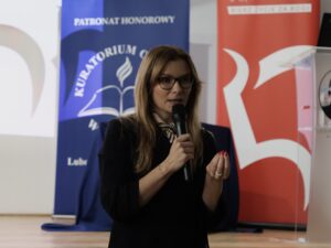 Młoda kobieta stoi na scenie i przemawia do publiczności, trzymając mikrofon w prawej dłoni. Ma długie blond włosy, nosi okulary o ciemnych oprawkach. Ubrana jest w elegancką czarną marynarkę i jasną apaszkę przewiązaną wokół szyi we wzór biało-czarnych pasków. Jej lewa ręka jest uniesiona i zgięta w geście podkreślającym sens wypowiedzi.

Tło stanowią pionowe roll-upy: granatowy z napisem „KURATORIUM…”, oraz czerwony z geometrycznym logotypem. Po lewej stronie widoczny jest fragment jasnego ekranu projekcyjnego.

Atmosfera zdjęcia sugeruje, że kobieta jest prelegentką prowadzącą wykład.