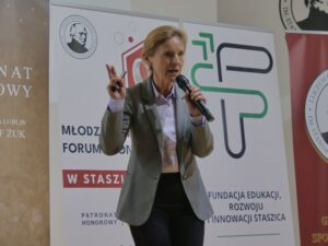 Dorosła kobieta – ta sama co na poprzednim zdjęciu – stoi na tle roll-upów i przemawia do publiczności. Jest uchwycona w dynamicznej pozie: w prawej ręce trzyma mikrofon przy ustach, a w lewej unosi dwa palce w geście przypominającym „dwa punkty” lub „peace”. Ubrana jest w szarą marynarkę, jasnofioletową koszulę i ciemne spodnie.

Za nią znajdują się trzy roll-upy: biały, brązowy i biało-zielony, z logotypami szkoły oraz fundacji edukacyjnej. Na jednym z nich widnieje znak graficzny przypominający literę „F” w nowoczesnym stylu.

Postawa kobiety jest dynamiczna, jakby wyjaśniała trudną koncepcję lub podkreślała ważny wkład edukacyjny.