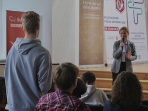 Ujęcie pokazuje salę wykładową z widownią młodzieży siedzącej w rzędach. Na pierwszym planie, odwrócony tyłem do aparatu, stoi młody chłopak w błękitnej bluzie z kapturem. Wygląda, jakby zadawał pytanie lub rozmawiał z prowadzącą.

Po prawej stronie kadru stoi dorosła kobieta prowadząca spotkanie – ubrana w szarą marynarkę i jasną koszulę. Trzyma mikrofon w prawej ręce i gestykuluje lewą, podkreślając wypowiedź. Wygląda przyjaźnie i z zaangażowaniem.

W tle znów widoczne są roll-upy w jasnych kolorach, przedstawiające logotypy wydarzenia edukacyjnego. Scena sprawia wrażenie żywej wymiany zdań.