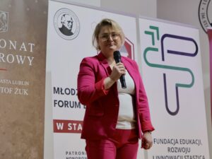 Dorosła kobieta stoi na scenie lub przed publicznością, trzymając mikrofon w prawej ręce. Ubrana jest w wyrazisty, różowy garnitur – marynarkę i spodnie – oraz białą bluzkę pod spodem. Kobieta ma krótkie blond włosy, okulary i poważny, ale życzliwy wyraz twarzy.

Tło stanowią trzy pionowe roll-upy z logotypami instytucji edukacyjnych i organizatorów wydarzenia. Na jednym z nich widoczne jest stylizowane logo przedstawiające profil mężczyzny, na innym geometryczne nowoczesne symbole. Kolorystyka roll-upów to biel, brąz, zieleń i czerwień.

Kobieta wygląda na osobę prowadzącą wykład lub przemówienie, stoi pewnie i spokojnie.