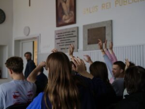 Zdjęcie przedstawia salę lekcyjną lub wykładową w jasnych barwach. W pierwszym planie widać tyły głów kilku siedzących uczniów. Uczniowie siedzą blisko siebie, patrząc w stronę prowadzącego. Kilkanaście rąk jest uniesionych w górę – uczniowie zgłaszają się do odpowiedzi lub chcą zabrać głos. Ręce są uniesione na różne wysokości, niektóre wyprostowane, inne lekko ugięte.

Po lewej stronie kadru w tle stoi nauczyciel, mężczyzna w okularach i ciemnej marynarce, lekko odwrócony bokiem. Na ścianie za uczniami widnieją duże tablice pamiątkowe z napisami oraz obraz religijny. Nad nimi znajduje się duży napis w języku łacińskim „SALUS REI PUBLICAE…”.

Całość przedstawia aktywną dyskusję, lekcję lub warsztat, podczas którego młodzież chętnie się angażuje.