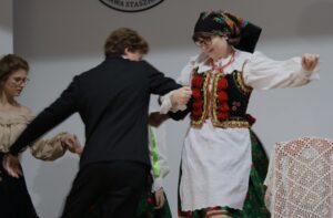 Na pierwszym planie para w tańcu: Dziewczyna: ubrana w tradycyjny, krakowski strój ludowy – czarny gorset bogato zdobiony koralikami i czerwonymi chwostami, biała koszula z długimi rękawami, biała zapaska z ażurowymi wycięciami, zielona spódnica z czerwonym wzorem kwiatowym. Na głowie czarna chusta z kolorowym, kwiecistym nadrukiem. Twarz pogodna, uśmiechnięta. Chłopak: ubrany w czarny garnitur, jasna koszula. Prowadzi dziewczynę w tańcu, łagodnie trzymając jej rękę. W tle widać innych tancerzy w ruchu, a także mebel nakryty białą, koronkową serwetą.