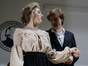 Ujęcie na dwie osoby: Dziewczyna w beżowej bluzce z długimi bufistymi rękawami oraz czarnej, długiej spódnicy. Włosy upięte, ma okulary. Twarz jest lekko odchylona, jakby śpiewała, mówiła lub była zamyślona. Chłopak w czarnym garniturze, biała koszula. Patrzy na dziewczynę z powagą i skupieniem. W tle fragment logo szkoły.