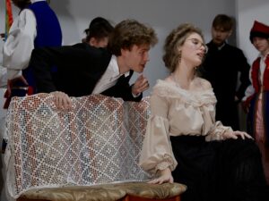 Dwie osoby siedzą na ławce przykrytej dużą, białą koronkową serwetą. Dziewczyna: w beżowej bluzce, z opuszczonymi ramionami, czarna długa spódnica. Siedzi w pozycji teatralnej, ręka oparta o ławkę, głowa lekko odchylona do tyłu, oczy półprzymknięte. Chłopak: pochylony w jej stronę, jakby szeptał coś lub rozmawiał. Ubrany w czarny garnitur. Za nimi stoją inni uczniowie – częściowo w strojach krakowskich, częściowo w ciemnych ubiorach stylizowanych na księdza czy dworzanina.