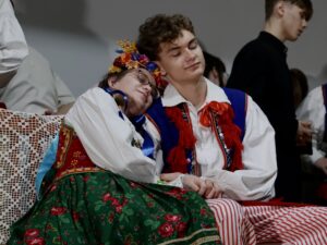 Na ławce z koronkową narzutą siedzi dwoje młodych ludzi: Dziewczyna w bogato zdobionym stroju krakowskim, na głowie ma wianek z dużą ilością kolorowych kwiatów (dominują pomarańcz, żółć, czerwień, niebieski). Ma białą koszulę, gorset w czerwono-zielonych barwach, zieloną spódnicę z czerwonymi kwiatami. Oparta o chłopaka, oczy ma zamknięte, wygląda jakby zasnęła. Chłopak w stroju krakowskim – biały kołnierzyk, pasiaki, granatowa kamizelka z czerwonymi chwostami. Też ma zamknięte oczy, oparty o nią policzkiem. W tle osoby w strojach galowych lub ludowych, tworzące spokojną atmosferę sceny.