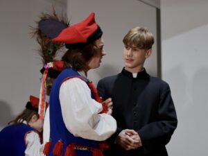 Po prawej stronie kadru stoi chłopak przebrany za księdza – czarna sutanna zapinana do samego pod szyję. Twarz pogodna, lekko uśmiechnięta. Po lewej stronie stoi druga osoba w stroju krakowskim – niebieska kamizelka, biała koszula, czerwona czapka z pawim piórem, czerwone chwosty przy gorsetowej kamizelce. Rozmawiają ze sobą – postać w stroju krakowskim mówi bardziej ekspresyjnie, gestykuluje rękami.