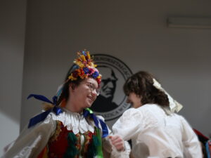 Dwie dziewczyny tańczą: Pierwsza, z lewej, ma na głowie ogromny, kolorowy wianek z kwiatów i długie wstążki w niebieskim, żółtym i białym kolorze. Ma uśmiechniętą twarz. Ubrana w tradycyjny strój krakowski: biała koszula, czerwony gorset, zielona spódnica w kwiaty. Druga, z prawej, ma białą bluzkę z długimi rękawami wykończonymi koronką i długą spódnicę. Włosy lekko falowane, związane z tyłu. Na ścianie w tle fragment logo szkoły.