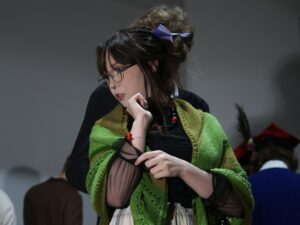 Na zdjęciu widać: Dziewczynę owiniętą zieloną, dzierganą chustą. Pod chustą ma czarną koszulę z półtransparentnymi rękawami. Włosy ma upięte, ozdobione jasnofioletową wstążką. Jej twarz jest spokojna, lekko zamyślona, a palce dłoni wykonują delikatny, teatralny gest. Za nią, nieco rozmyte, widać inne osoby w ciemniejszych strojach.