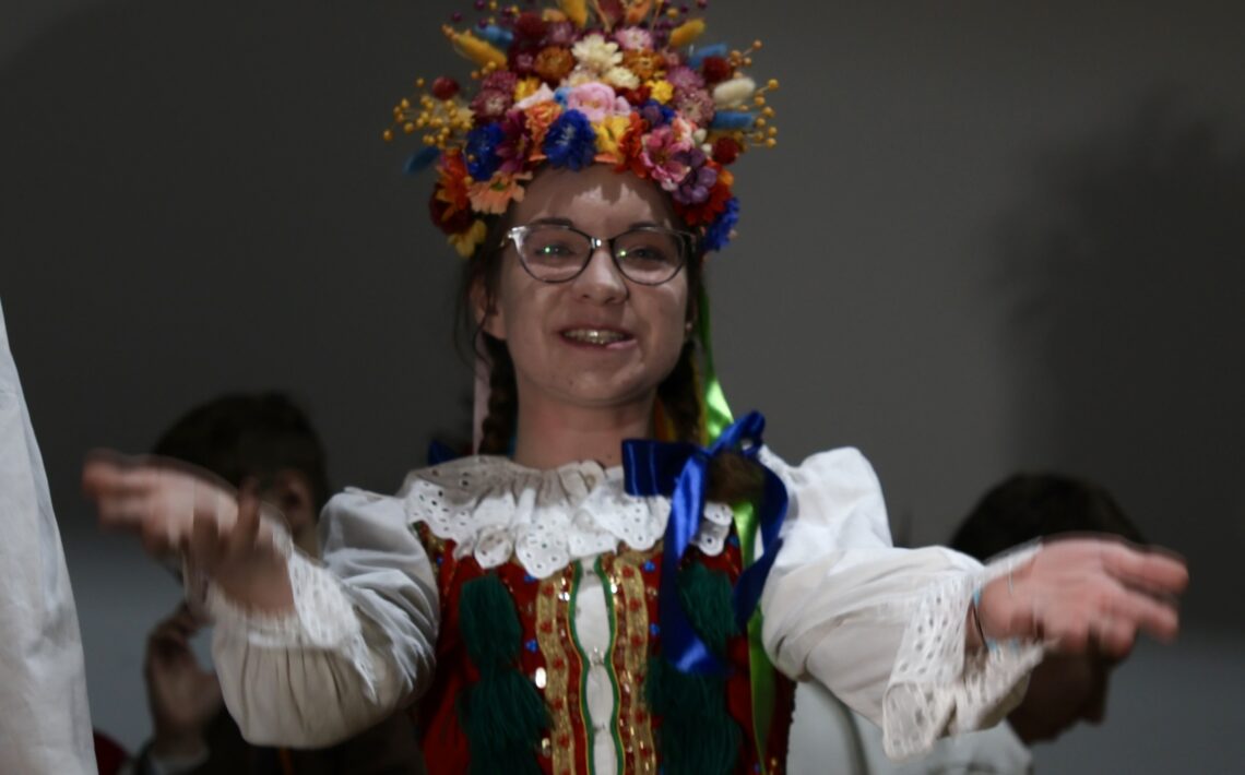 W kadrze znajduje się uśmiechnięta młoda aktorka w pełnym, bogatym stroju krakowskim: Na głowie ma duży, kolorowy wianek z kwiatów w odcieniach żółci, czerwieni, niebieskiego, różu i pomarańczu. Włosy splecione w warkocze. Gorset czerwony, bogato zdobiony, z charakterystycznymi zielonymi chwostami. Biała koszula z koronkowymi rękawami. Ręce wyciąga do przodu w geście powitalnym lub tanecznym, jakby zapraszała do zabawy.