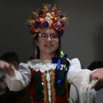 W kadrze znajduje się uśmiechnięta młoda aktorka w pełnym, bogatym stroju krakowskim: Na głowie ma duży, kolorowy wianek z kwiatów w odcieniach żółci, czerwieni, niebieskiego, różu i pomarańczu. Włosy splecione w warkocze. Gorset czerwony, bogato zdobiony, z charakterystycznymi zielonymi chwostami. Biała koszula z koronkowymi rękawami. Ręce wyciąga do przodu w geście powitalnym lub tanecznym, jakby zapraszała do zabawy.