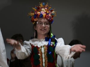 W kadrze znajduje się uśmiechnięta młoda aktorka w pełnym, bogatym stroju krakowskim: Na głowie ma duży, kolorowy wianek z kwiatów w odcieniach żółci, czerwieni, niebieskiego, różu i pomarańczu. Włosy splecione w warkocze. Gorset czerwony, bogato zdobiony, z charakterystycznymi zielonymi chwostami. Biała koszula z koronkowymi rękawami. Ręce wyciąga do przodu w geście powitalnym lub tanecznym, jakby zapraszała do zabawy.