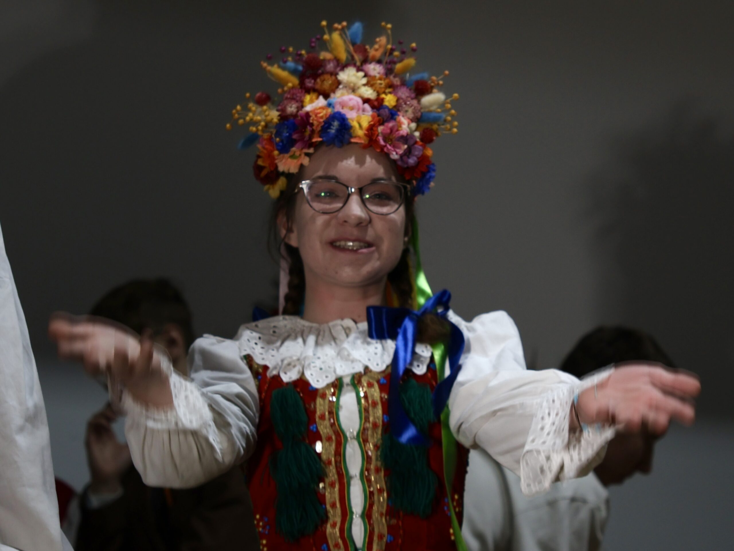 W kadrze znajduje się uśmiechnięta młoda aktorka w pełnym, bogatym stroju krakowskim: Na głowie ma duży, kolorowy wianek z kwiatów w odcieniach żółci, czerwieni, niebieskiego, różu i pomarańczu. Włosy splecione w warkocze. Gorset czerwony, bogato zdobiony, z charakterystycznymi zielonymi chwostami. Biała koszula z koronkowymi rękawami. Ręce wyciąga do przodu w geście powitalnym lub tanecznym, jakby zapraszała do zabawy.