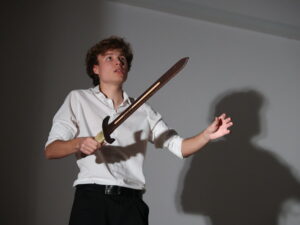 Na zdjęciu widać młodego aktora stojącego w białej koszuli i czarnych spodniach: Trzyma drewniany miecz obiema rękami, jeden koniec zwrócony lekko ku górze. Twarz ma uniesioną, spojrzenie skierowane w górę, wyraz poważny i pełen napięcia. Jego cień na ścianie powiększa sylwetkę, dodając dramatyzmu całej scenie.