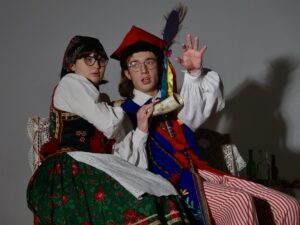 Na zdjęciu widać dwoje młodych aktorów siedzących blisko siebie na ławce przykrytej białą, koronkową narzutą. Oboje mają na sobie tradycyjne stroje ludowe z regionu krakowskiego. Postacie i ich ustawienie Dziewczyna po lewej: Ma czarną chustę w kwiaty zawiązaną na głowie, okulary, białą koszulę z koronkowym kołnierzem i bogato zdobiony ciemny gorset z czerwonymi frędzlami. Spódnica zielona w czerwone róże. Jej ciałem delikatnie przylega do chłopaka, jakby była czymś zaskoczona lub przestraszona. Patrzy lekko w bok, oczy szeroko otwarte. Chłopak po prawej: Ubrany w strój krakowski: biała koszula z bufiastymi rękawami, niebieska kamizelka zdobiona czerwonymi chwostami, czerwono-biało pasiakowe spodnie oraz tradycyjna czerwona czapka z długimi kolorowymi wstążkami i pawim piórem. W rękach trzyma róg pasterski, wykonany z jasnego materiału. Lewą rękę unosi w górę w geście ostrzegawczym, jakby chciał uciszyć publiczność lub wskazać niebezpieczeństwo. Twarz ma napiętą, usta lekko rozchylone, jakby coś mówił. Tło jest szare, jednolite. Po prawej stronie widoczne są butelki i zastawa na stole, częściowo przysłonięte cieniem.