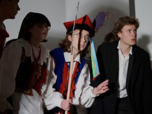 W kadrze znajdują się trzy osoby, wszystkie pochylone do przodu, jakby nasłuchiwały lub obserwowały coś niepokojącego. Osoby: Dziewczyna po lewej: W stroju krakowskim (ciemny gorset z czerwonymi frędzlami, biała koszula z koronką). Na głowie ma czarną chustę w kwiaty. Twarz poważna, usta lekko rozchylone. Chłopak w środku: W pełnym stroju krakowskim: niebieska kamizelka, czerwone chwosty, czapka krakowska z pawim piórem i kolorowymi wstążkami. W ręku trzyma długi kij lub tyczkę. Oczy szeroko otwarte, kierunek patrzenia na coś poza kadrem, twarz skupiona. Chłopak po prawej: Ubrany w czarny garnitur i białą koszulę. Jego wyraz twarzy jest również poważny; pochyla się do przodu, jakby chciał lepiej zobaczyć. Tło stanowią gładkie ściany szkoły, a światło punktowe rzuca ostre cienie postaci na ścianę.