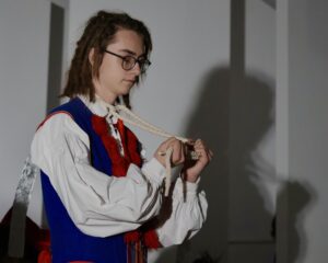 Na zdjęciu znajduje się młody aktor w stroju krakowskim, ujęty z profilu. Wygląd postaci Na głowie nie ma czapki, widoczne są włosy w formie dreadów. Twarz spokojna, skupiona. Ubrany w tradycyjny niebieski kaftan z czerwonymi chwostami oraz białą, szeroką koszulę z bufiastymi rękawami. W dłoniach trzyma grubą lnianą linę, uformowaną w pętlę. Lina opiera się na jego szyi, ale nie wygląda, jakby sprawiała mu ból – gest jest raczej teatralny, symboliczny. Cień jego sylwetki pada wyraźnie na ścianę za nim, tworząc dramatyczny efekt.