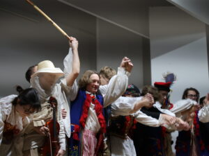 Zdjęcie przedstawia grupę kilkunastu aktorów, stojących w szeregu podczas finałowego ukłonu przedstawienia. Najważniejsze szczegóły: Po lewej stronie osoba grająca skrzypce, w jasnym stroju i szerokim, słomkowym kapeluszu z woalem, unosi w górę kij lub bat. Obok niej stoją osoby w strojach ludowych (niebieskie kamizelki, czerwone chwosty, pasiaki). Wszystkie osoby trzymają się za ręce, unosząc je wysoko, tworząc łuk nad głowami. Wszyscy pochylają się lekko do przodu. W tle wyraźnie widoczne światło teatralne.