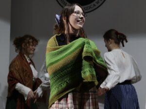 Na pierwszym planie widać uśmiechniętą dziewczynę: Ma na sobie zielono-żółtą, grubą, dzierganą chustę zarzuconą na ramiona. Pod nią czarną bluzkę z półprzezroczystymi rękawami i długą spódnicę z wielokolorowej kraty. Na głowie upięte włosy, przewiązane fioletową wstążką. W tle dwie kolejne postacie tańczą – jedna w brązowej chuście i białej koszuli, druga tyłem, w białej bluzce i granatowej spódnicy.