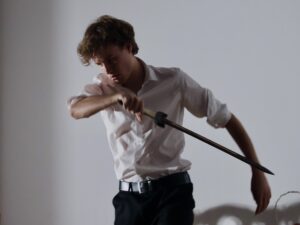 Zdjęcie przedstawia młodego aktora wykonującego intensywny ruch: Ma na sobie białą koszulę z podwiniętymi rękawami i czarne spodnie. W prawej ręce trzyma drewniany miecz, uniesiony skośnie. Jego ciało jest skręcone, jakby wykonywał zamach lub przygotowywał się do obrony. Twarz poważna, lekko zmęczona, brwi ściągnięte. Cień postaci tworzy wyraźny, powiększony kształt na ścianie, co wzmacnia dramatyzm sceny.