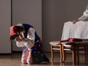 W centrum zdjęcia znajduje się młody aktor: Klęczy na scenie, pochylony nisko, niemal dotykając czołem rąk. Ubrany w niebieską kamizelkę z czerwonymi frędzlami, białą koszulę i czerwono-białe pasiaki. Na głowie ma czerwoną czapkę krakowską z pawim piórem. Za nim widać ławkę przykrytą koronkową narzutą oraz różowe i czerwone elementy kostiumów. Oświetlenie jest punktowe – postać znajduje się w jasnym kręgu światła, reszta sceny w półcieniu.