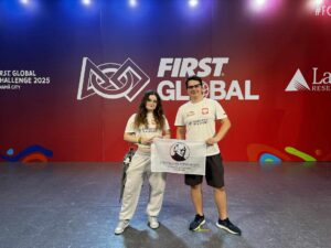 Na scenie z kolorowym tłem stoją dwie młode osoby — dziewczyna i chłopiec — oboje w białych koszulkach z napisem „ROBOTICS IS A SPORT” oraz godłem Polski.
Tło sceny wypełnia duże logo zawodów „FIRST GLOBAL CHALLENGE 2025 PANAMA CITY”.
Dziewczyna po lewej stronie ma długie, falowane włosy, okulary i szare spodnie dresowe. Chłopiec po prawej stronie ma krótkie włosy i czarne spodenki.
Oboje trzymają flagę szkoły z portretem Stefana Żeromskiego.
Światło sceniczne podkreśla ich sylwetki, tło ma odcienie czerwieni, fioletu i błękitu — nadając zdjęciu oficjalny, uroczysty charakter.
