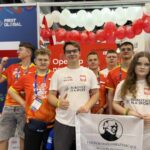 Sześć młodych osób stoi razem przed stoiskiem drużyny narodowej. Czterech chłopców i jedna dziewczyna mają na sobie pomarańczowo-czerwone koszulki drużynowe, a druga dziewczyna — białą koszulkę z napisem „ROBOTICS IS A SPORT”. Osoba z przodu po prawej stronie trzyma biało-czarną flagę z portretem Stefana Żeromskiego i napisem „Liceum Ogólnokształcące im. Stefana Żeromskiego w Strzelinie”. W tle widoczna jest ściana ozdobiona biało-czerwonymi balonami oraz logo zawodów „FIRST GLOBAL”. Po lewej stronie widać fragment flagi Polski. Uczestnicy pozują do wspólnego zdjęcia, część z nich pokazuje kciuki w górę, atmosfera jest radosna i pełna dumy.