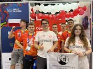 Sześć młodych osób stoi razem przed stoiskiem drużyny narodowej.
Czterech chłopców i jedna dziewczyna mają na sobie pomarańczowo-czerwone koszulki drużynowe, a druga dziewczyna — białą koszulkę z napisem „ROBOTICS IS A SPORT”.
Osoba z przodu po prawej stronie trzyma biało-czarną flagę z portretem Stefana Żeromskiego i napisem „Liceum Ogólnokształcące im. Stefana Żeromskiego w Strzelinie”.
W tle widoczna jest ściana ozdobiona biało-czerwonymi balonami oraz logo zawodów „FIRST GLOBAL”. Po lewej stronie widać fragment flagi Polski.
Uczestnicy pozują do wspólnego zdjęcia, część z nich pokazuje kciuki w górę, atmosfera jest radosna i pełna dumy.