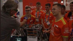 Grupa młodych chłopców stoi wokół robota zbudowanego z metalu i kabli.
Wszyscy mają pomarańczowo-czerwone koszulki z napisem „TEAM POLAND” i logotypami „FIRST GLOBAL” oraz sponsorów.
Przed nimi stoi starszy mężczyzna w szarej koszulce, trzymający mikrofon — wygląda na dziennikarza przeprowadzającego wywiad.
Chłopiec w środku odpowiada na pytanie, uśmiechając się, a obok stoją jego koledzy z identyfikatorami na niebieskich smyczach.
Na pierwszym planie widać tabliczkę z napisem „TEAM POLAND”, a w tle czerwone tło stoiska z nazwami partnerów drużyny.
