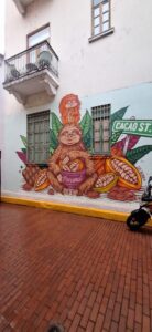 Fotografia przedstawia mural na jasnej ścianie budynku z dwoma oknami zabezpieczonymi kratami.
Na murze znajduje się barwne malowidło dużego, brązowego leniwca siedzącego pośrodku kompozycji. Zwierzę trzyma w łapach różową miseczkę z napisem „Panama”.
Za leniwcem rosną duże, zielone liście kakaowca oraz owoce kakaowca w przekroju, pokazujące nasiona.
Na głowie leniwca siedzi mniejsze zwierzątko — również leniwiec, pomarańczowy, uśmiechnięty.
Po prawej stronie muralu widnieje zielony napis „Cacao St.” (czyli „Ulica Kakao”).
Przed ścianą biegnie wąski chodnik z czerwonej cegły, a po prawej stronie kadru widoczny jest fragment roweru lub skutera.