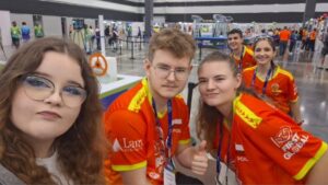 Na zdjęciu widać pięć młodych osób w dużej hali, prawdopodobnie podczas międzynarodowych zawodów robotycznych.
Z przodu po lewej stronie stoi dziewczyna z długimi, falowanymi włosami, w okrągłych okularach, ubrana w jasną koszulkę. Robi selfie i delikatnie się uśmiecha.
Obok niej, po prawej stronie, stoi trójka młodych ludzi w pomarańczowo-czerwonych koszulkach drużynowych z napisem „POLAND” i logo „FIRST GLOBAL”. Dwóch chłopców i jedna dziewczyna — chłopiec z blond włosami w okularach pokazuje kciuk do góry, dziewczyna obok niego również pokazuje gest „okej”.
W tle widać duże, otwarte pomieszczenie z wieloma stanowiskami i innymi uczestnikami w kolorowych strojach — panuje atmosfera zawodów.