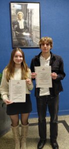 Na zdjęciu stoją dwie młode osoby – dziewczyna po lewej i chłopak po prawej – na tle niebieskiej ściany. Oboje trzymają przed sobą kartki w przezroczystych koszulkach z oficjalnym dokumentem zatytułowanym „ZAŚWIADCZENIE” (tekst w języku polskim, z godłem i nagłówkiem). Dziewczyna ma jasne, długie włosy, jasny sweter i ciemną spódnicę; chłopak ma jasnobrązowe włosy, białą koszulkę i ciemną kurtkę. Nad nimi wisi oprawiony portret Marii Skłodowskiej-Curie. Zdjęcie ma charakter pamiątkowy, dokumentujący otrzymanie zaświadczeń.