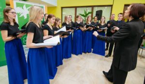 Młodzieżowy chór podczas występu w nowoczesnym, jasnym wnętrzu. Śpiewające osoby stoją półkolem, trzymają czarne teczki z nutami. Kobiety ubrane są w czarne bluzki i długie, niebieskie spódnice, mężczyźni w czarne stroje z niebieskimi akcentami. Przed zespołem stoi dyrygentka, zwrócona twarzą do chóru, z uniesionymi rękami. W tle zielono-żółte ściany, roślina doniczkowa i elementy wyposażenia budynku.