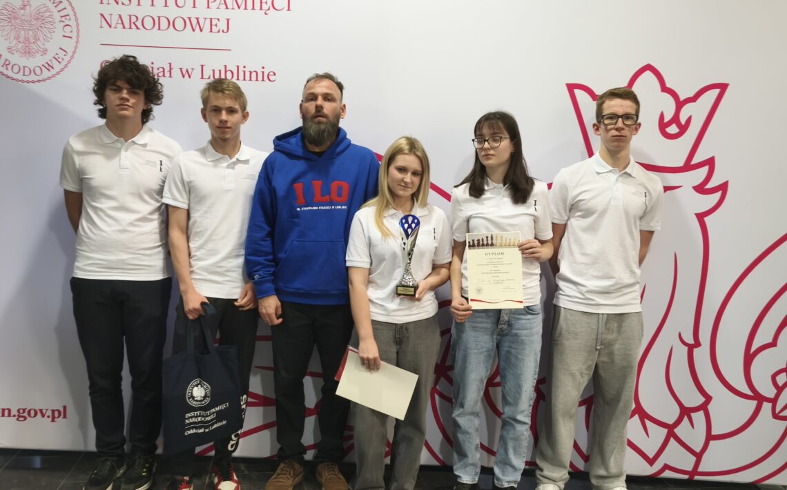 Grupa siedmiu młodych osób stoi przed ścianką z logo Instytutu Pamięci Narodowej w Lublinie. Uczestnicy są ubrani sportowo, część trzyma dyplomy i nagrody, wszyscy pozują do wspólnego zdjęcia.