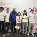 Grupa siedmiu młodych osób stoi przed ścianką z logo Instytutu Pamięci Narodowej w Lublinie. Uczestnicy są ubrani sportowo, część trzyma dyplomy i nagrody, wszyscy pozują do wspólnego zdjęcia.