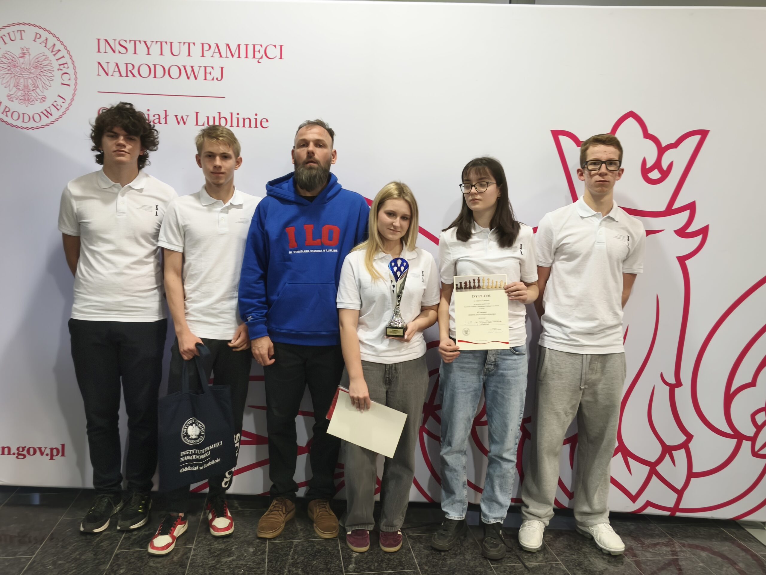 Grupa siedmiu młodych osób stoi przed ścianką z logo Instytutu Pamięci Narodowej w Lublinie. Uczestnicy są ubrani sportowo, część trzyma dyplomy i nagrody, wszyscy pozują do wspólnego zdjęcia.