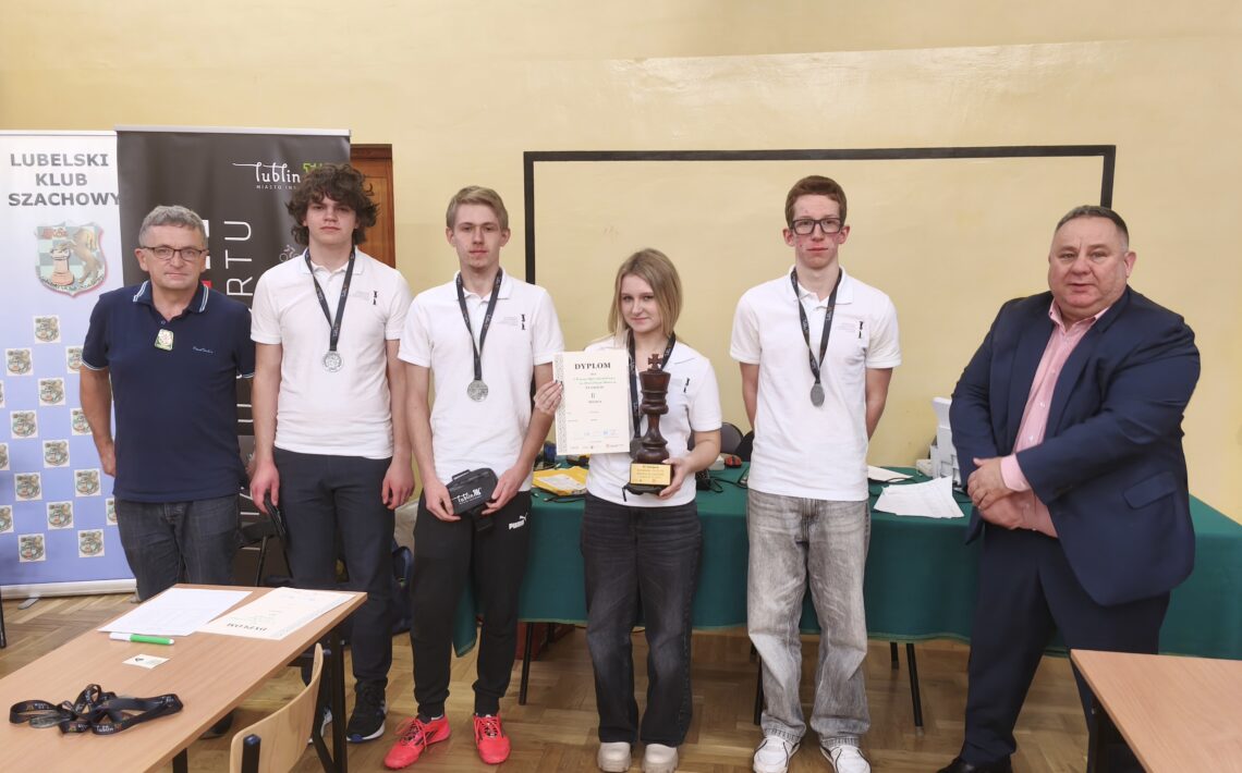 Grupa sześciu młodych osób z medalami na szyi stoi w sali lekcyjnej obok dwóch dorosłych mężczyzn. Na stole przed nimi leżą dokumenty i sprzęt, wszyscy pozują do pamiątkowego zdjęcia.