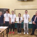 Grupa sześciu młodych osób z medalami na szyi stoi w sali lekcyjnej obok dwóch dorosłych mężczyzn. Na stole przed nimi leżą dokumenty i sprzęt, wszyscy pozują do pamiątkowego zdjęcia.