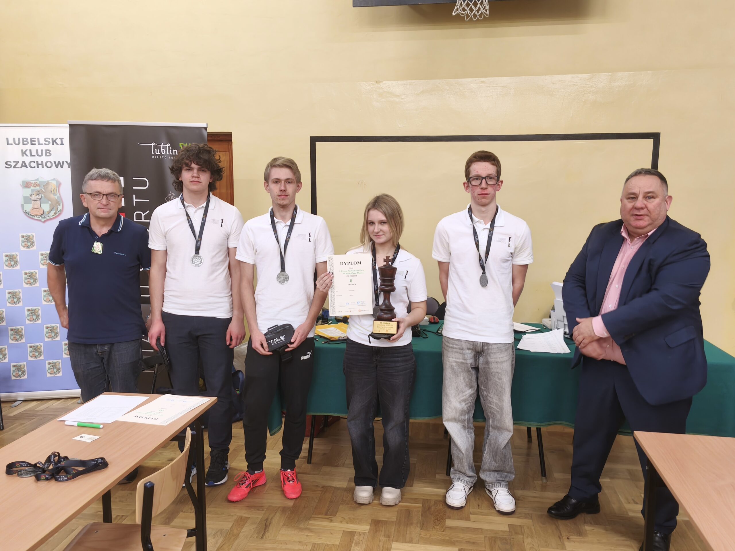 Grupa sześciu młodych osób z medalami na szyi stoi w sali lekcyjnej obok dwóch dorosłych mężczyzn. Na stole przed nimi leżą dokumenty i sprzęt, wszyscy pozują do pamiątkowego zdjęcia.