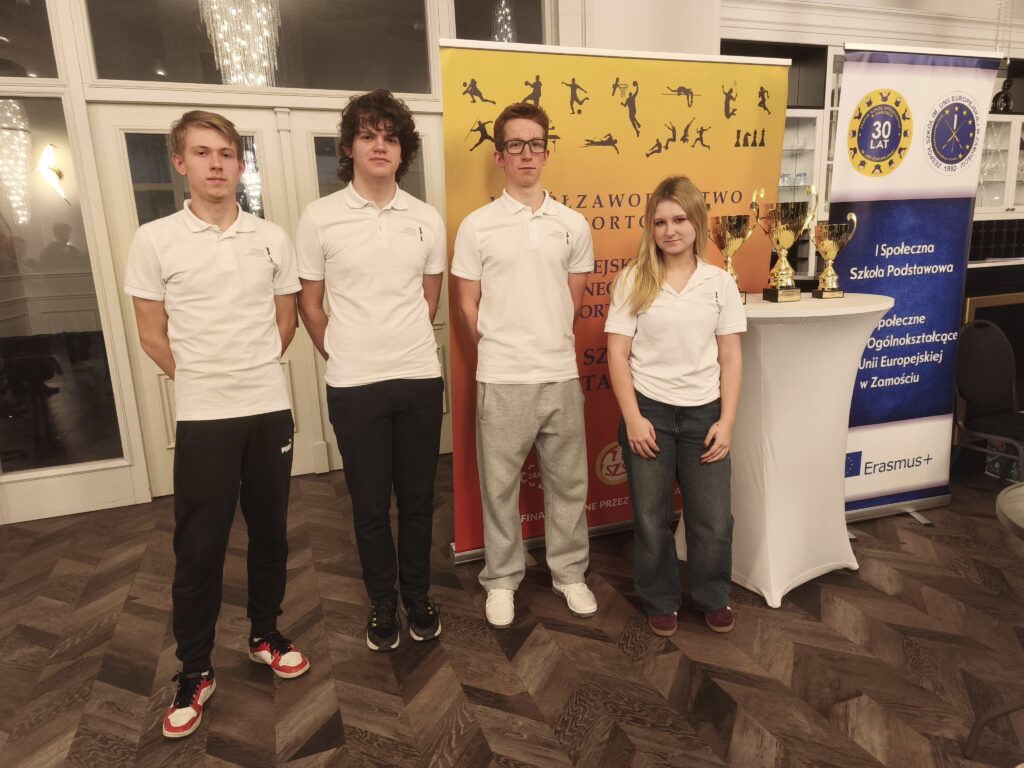 Czworo młodych osób i jedna osoba dorosła stoją w holu przed żółtą ścianką informacyjną oraz roll-upem. Uczestnicy są ubrani na jasno, pozują do wspólnego zdjęcia podczas wydarzenia lub konkursu.