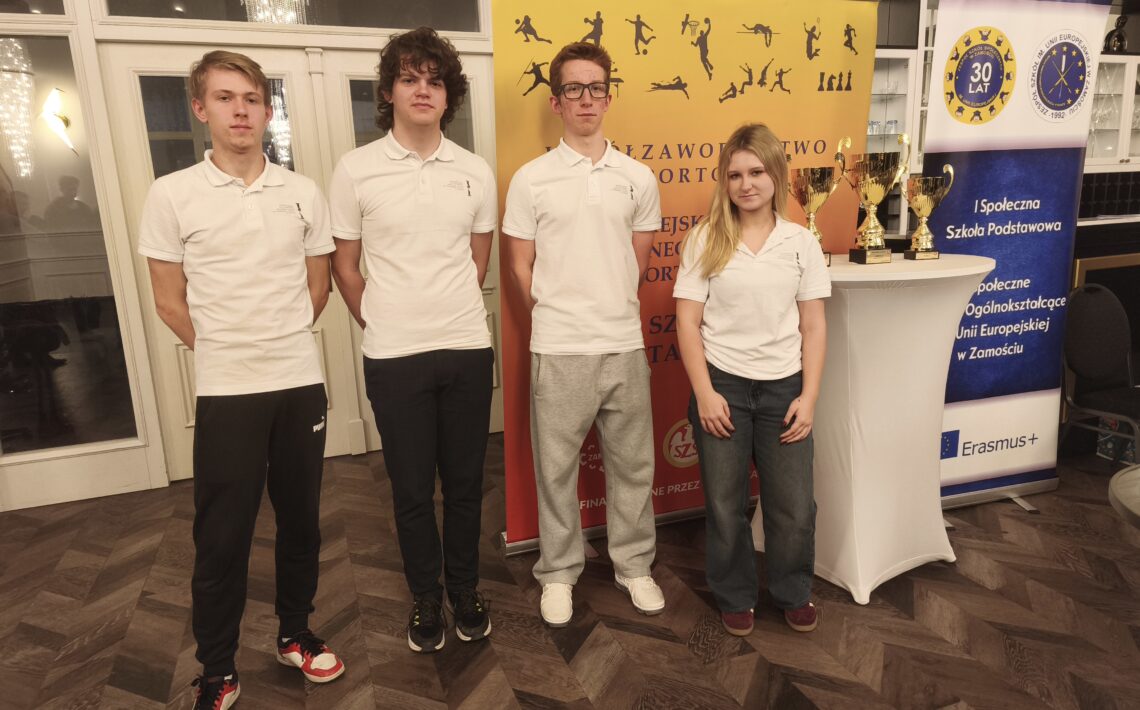 Czworo młodych osób i jedna osoba dorosła stoją w holu przed żółtą ścianką informacyjną oraz roll-upem. Uczestnicy są ubrani na jasno, pozują do wspólnego zdjęcia podczas wydarzenia lub konkursu.