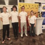 Czworo młodych osób i jedna osoba dorosła stoją w holu przed żółtą ścianką informacyjną oraz roll-upem. Uczestnicy są ubrani na jasno, pozują do wspólnego zdjęcia podczas wydarzenia lub konkursu.