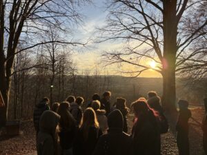 Grupa młodych ludzi stoi na zewnątrz na wzgórzu, patrząc na zachód słońca nad krajobrazem drzew i pól.