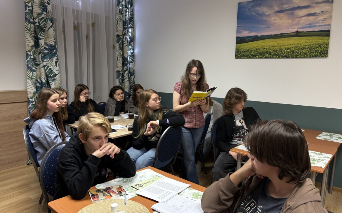 Młodzież siedzi przy stołach w sali i uczestniczy w zajęciach; jedna z uczennic czyta książkę na głos, pozostali słuchają.