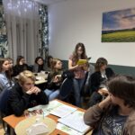 Młodzież siedzi przy stołach w sali i uczestniczy w zajęciach; jedna z uczennic czyta książkę na głos, pozostali słuchają.