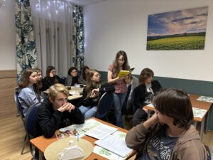 Młodzież siedzi przy stołach w sali i uczestniczy w zajęciach; jedna z uczennic czyta książkę na głos, pozostali słuchają.