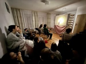 Grupa młodych osób siedzi w pokoju i ogląda teatr cieni wyświetlany na prowizorycznym ekranie; panuje kameralna, skupiona atmosfera.