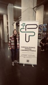 Na zdjęciu widać wnętrze nowoczesnego, betonowego budynku z wysokim sufitem i liniowymi lampami LED. Podłoga jest ciemna i błyszcząca. Po lewej stronie fotografii stoi osoba – dorosła kobieta o jasnych włosach do ramion, uśmiechnięta, ubrana w koszulę w czerwono-białą kratę, czarną koszulkę i ciemne dżinsy. Ma na nogach czarno-białe sportowe buty.

Obok kobiety stoi pionowy biały roll-up z dużym logo składającym się z dwóch stylizowanych strzałek i linii układających się w abstrakcyjny znak w kolorach zielonym i czarnym. Pod logo widnieje nazwa organizacji:
„Fundacja Edukacji, Rozwoju i Innowacji Staszica”.

Poniżej znajdują się dane kontaktowe:
Adres: Al. Racławickie 26, 20-043 Lublin
NIP: 7123495882
REGON: 542134643
Telefon: +48 789 478 389
E-mail: kontakt@fundacjastaszica.pl

Strona: www.fundacjastaszica.pl

Na dole widoczny jest kod QR.

W tle widać betonowe filary oraz tablice z afiszami i plakatami. Po prawej stronie siedzą dwie młode osoby, zajęte rozmową i telefonem.